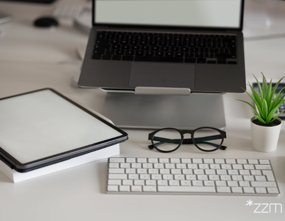 Nowoczesne stanowisko pracy na białym biurku. Na podwyższeniu stoi otwarty laptop z jasnym, pustym ekranem, a przed nim leżą biała klawiatura i czarne okulary. Po lewej stronie znajduje się tablet oparty na książce, a po prawej niewielka zielona roślina w białej doniczce oraz szklanka z wodą.