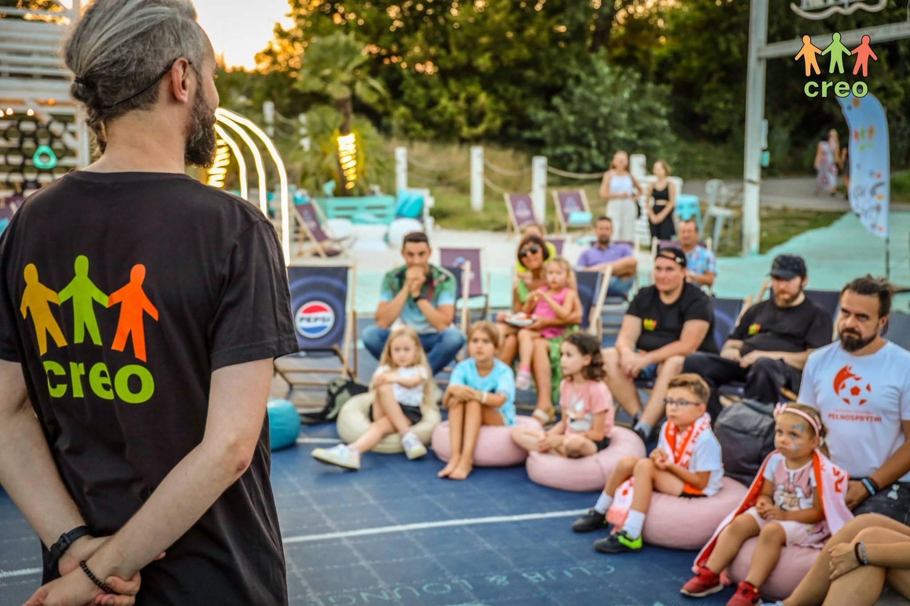 Osoba w koszulce z napisem CREO stoi przed słuchacvzami. Sa nimi rodziny z dziećmi, wszyscy siedza na plaży.