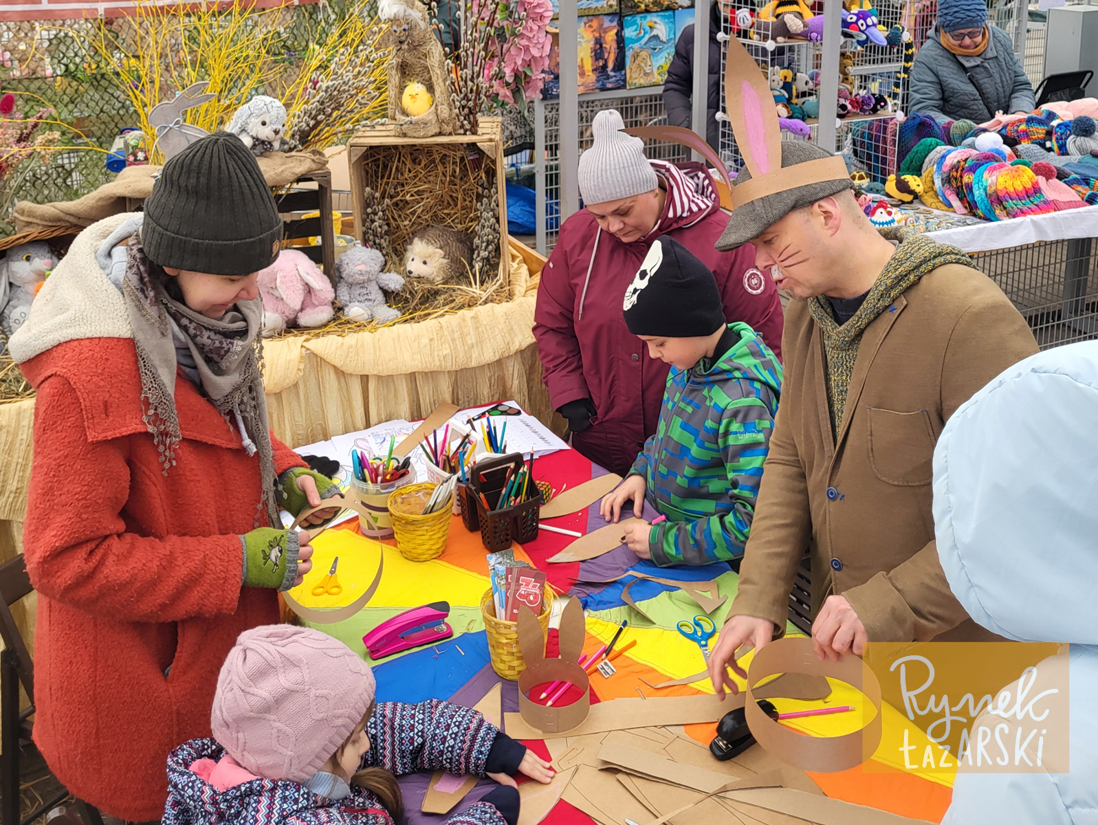 Osoby w różnym wielu stoją przu stole i wykonują prace plastyczne i ozdoby świąteczne.