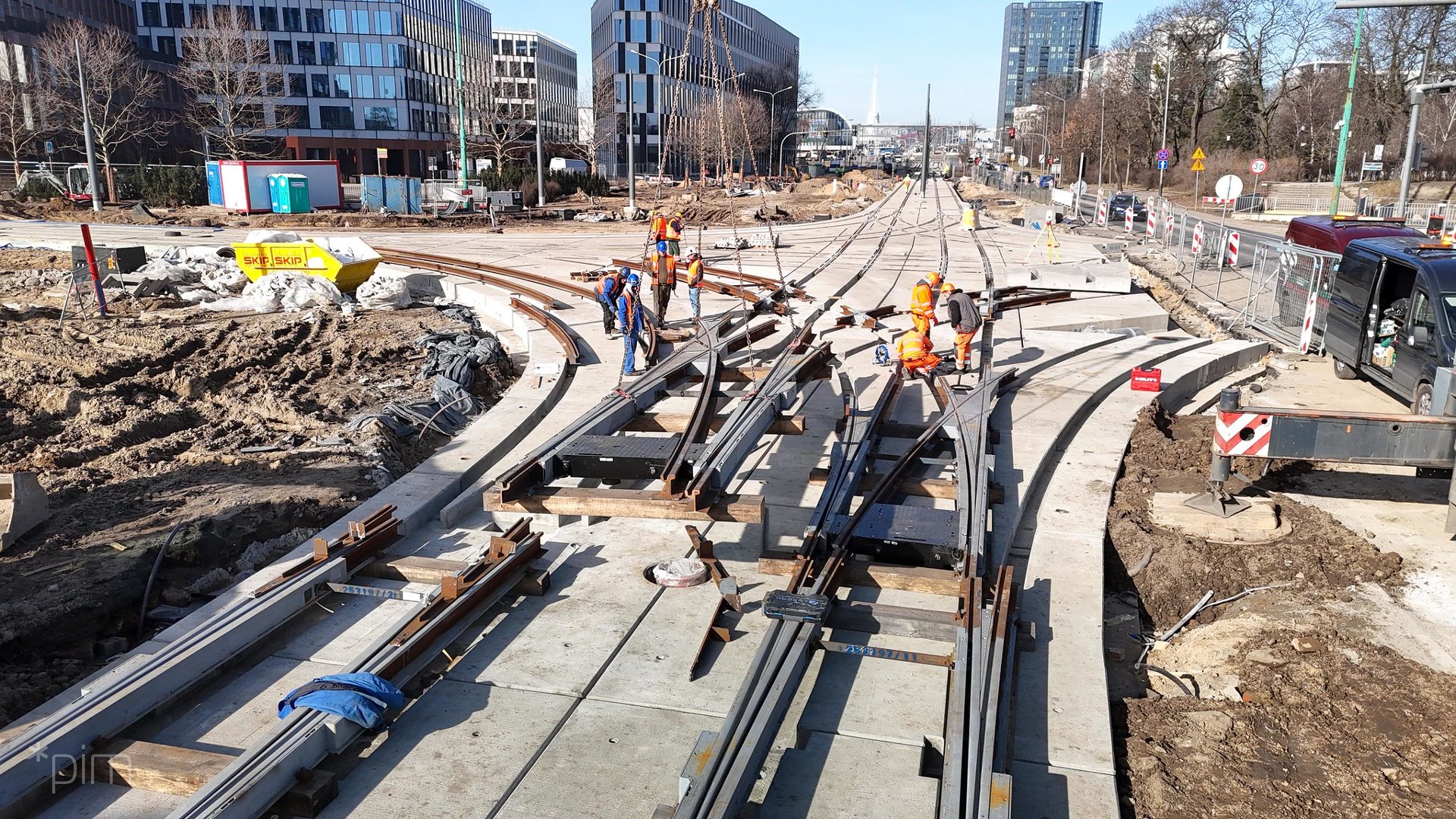Plac budowy , na którym widać elementy torowiska tramwajowego. Przy nim widac pracowników ubranych w stroje robocze w kaskach. W tle widać wysokie budynki - biurowce.