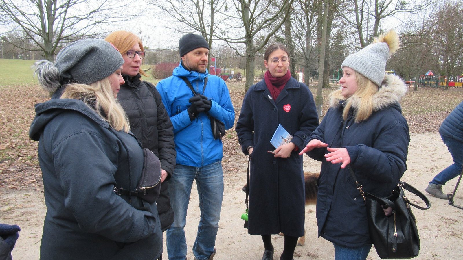 Zdjęcie przedstawia grupkę ludzi w parku. Jedna z kobiet gestykuluje, pozostali ją słuchają.
