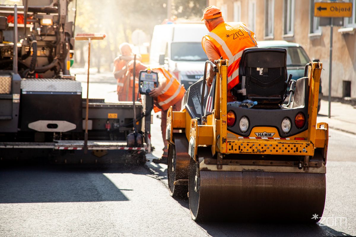 Robotnicy drogowi w pomarańczowych kamizelkach pracują na ulicy. Jeden z nich siedzi na walcu drogowym, a drugi obsługuje maszynę do układania asfaltu. W tle widać białą furgonetkę i znaki drogowe.