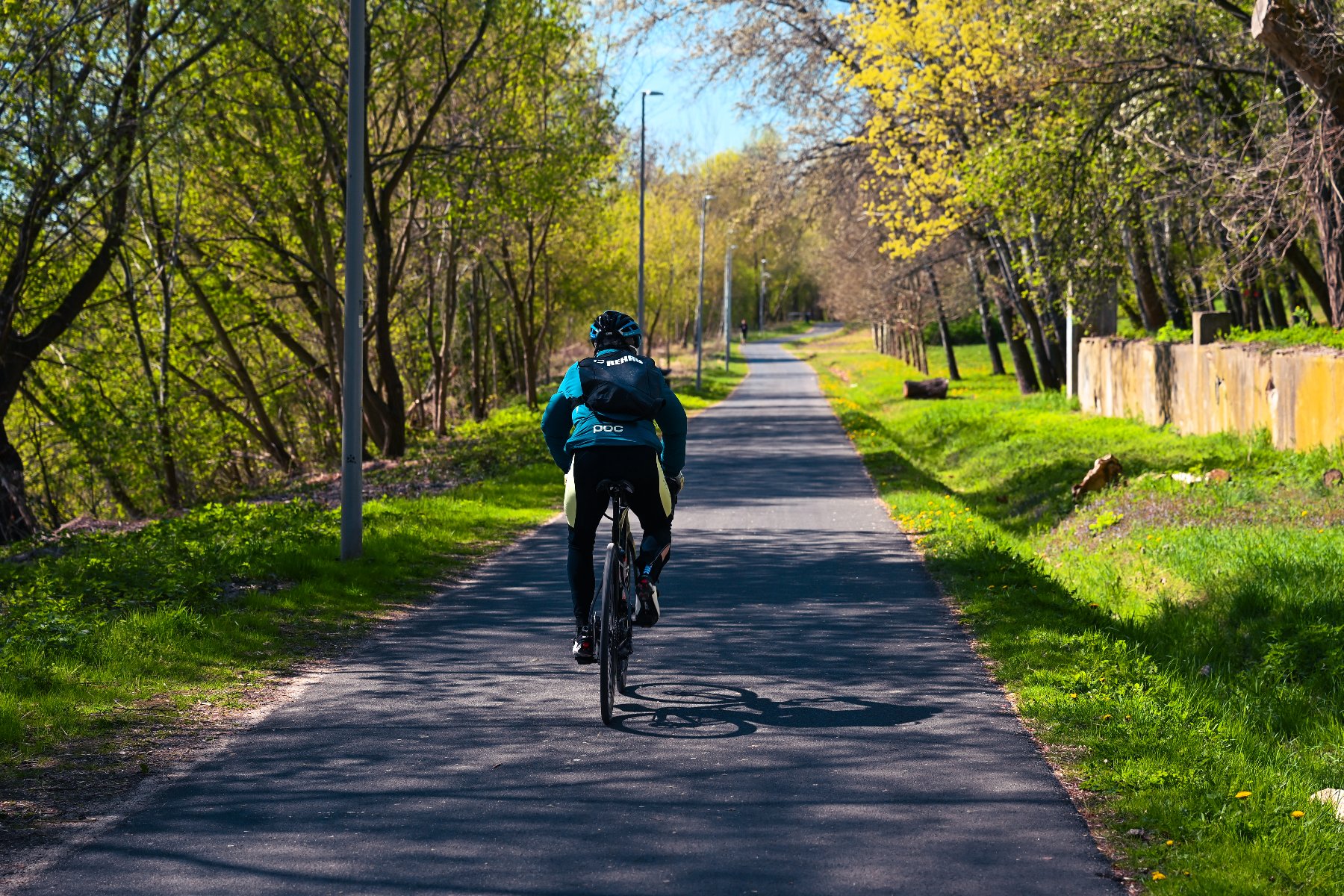 Rowerzysta w kasku i sportowym stroju jedzie asfaltową ścieżką pośród zielonych drzew i trawników.
