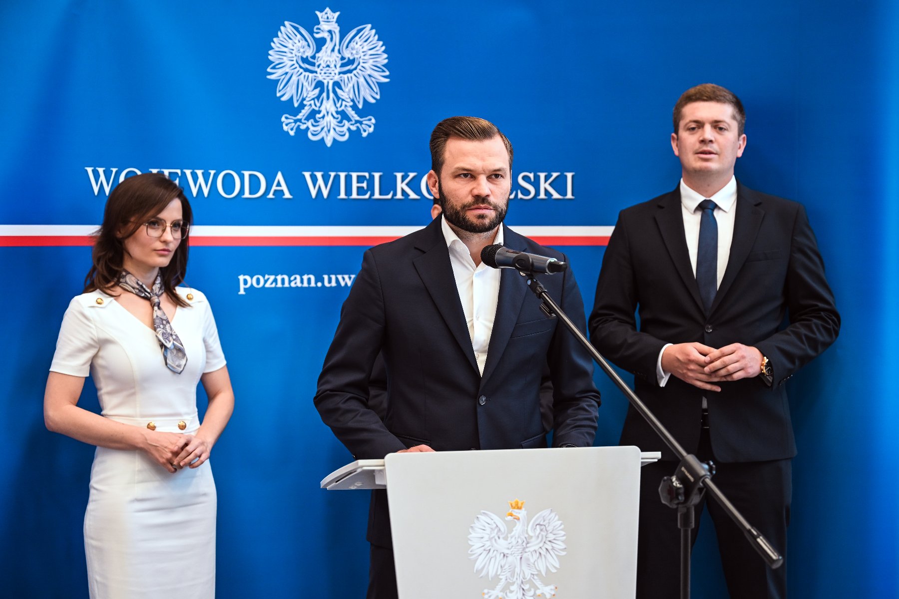 rzy osoby w formalnych strojach stoją na tle niebieskiego baneru z napisem "WOJEWODA WIELKOPOLSKI". Mężczyzna w centrum przemawia do mikrofonu, a kobieta w białej sukience stoi po jego lewej stronie. Na mównicy znajduje się godło.