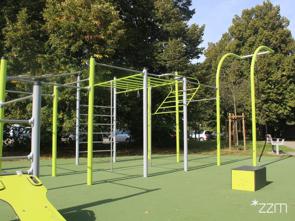 Strefa street workout z zielono-szarymi drążkami, drabinkami i kółkami do ćwiczeń na zielonej nawierzchni amortyzującej upadki.
