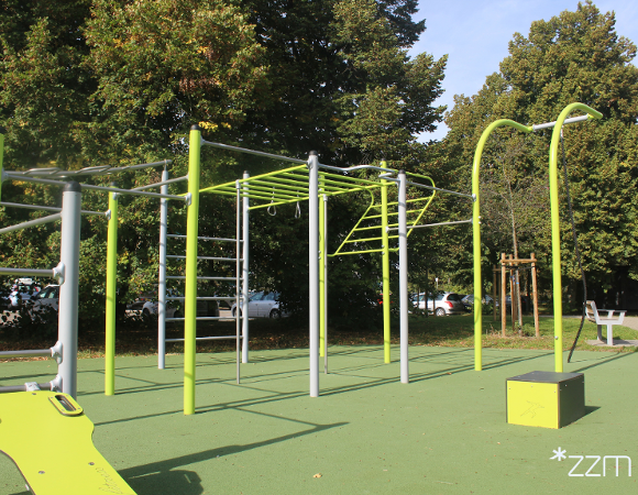 Strefa street workout z zielono-szarymi drążkami, drabinkami i kółkami do ćwiczeń na zielonej nawierzchni amortyzującej upadki.