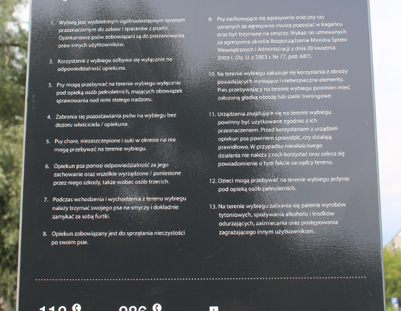 Tablica informacyjna z napisem "Wybieg dla psów" oraz symbolem psa. Poniżej znajduje się szczegółowy, trzynastopunktowy regulamin korzystania z wybiegu, a na dole numery alarmowe 112 i 986 oraz dane administratora terenu.