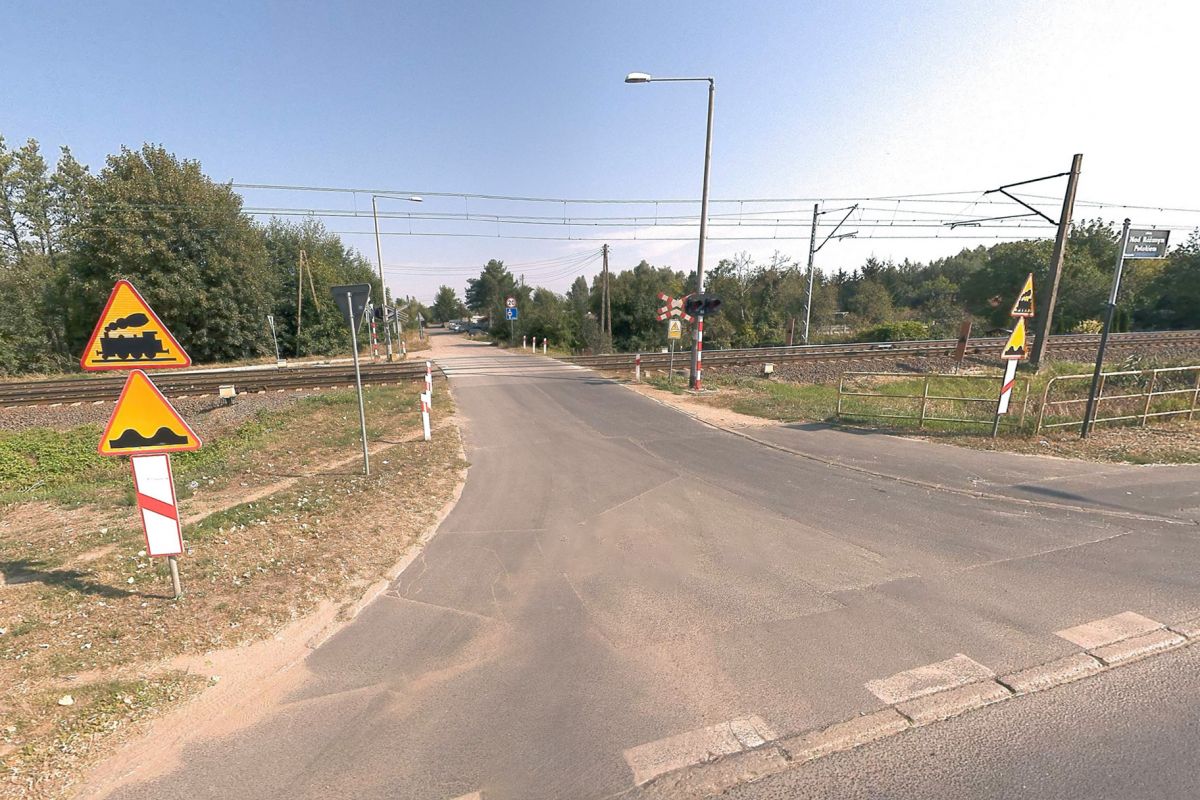 Tory kolejowe przecinają drogę asfaltową. Przy drodze stoją znaki ostrzegawcze, sygnalizacja świetlna oraz lampy uliczne. W tle widać drzewa, zarośla i kawałek nieba.