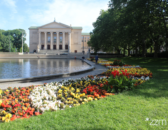 Widok na fontannę w parku Adama Mickiewicza i otaczające ją wielobarwne rabaty. W tle znajduje się gmach Teatru Wielkiego.