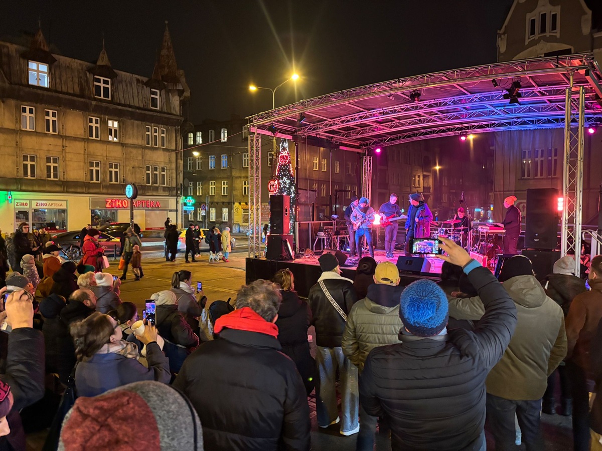 Widok na kolorowo podświetloną scenę. Przed nią stoją widzowie. Na scenie stoją muzycy i grają na gitarach.