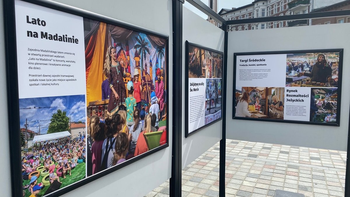 Widok na zdjęcia z wystawy umieszczone w sztalugach. Na głownym planie widok na fotografię z napisem "Lato na Madalinie"