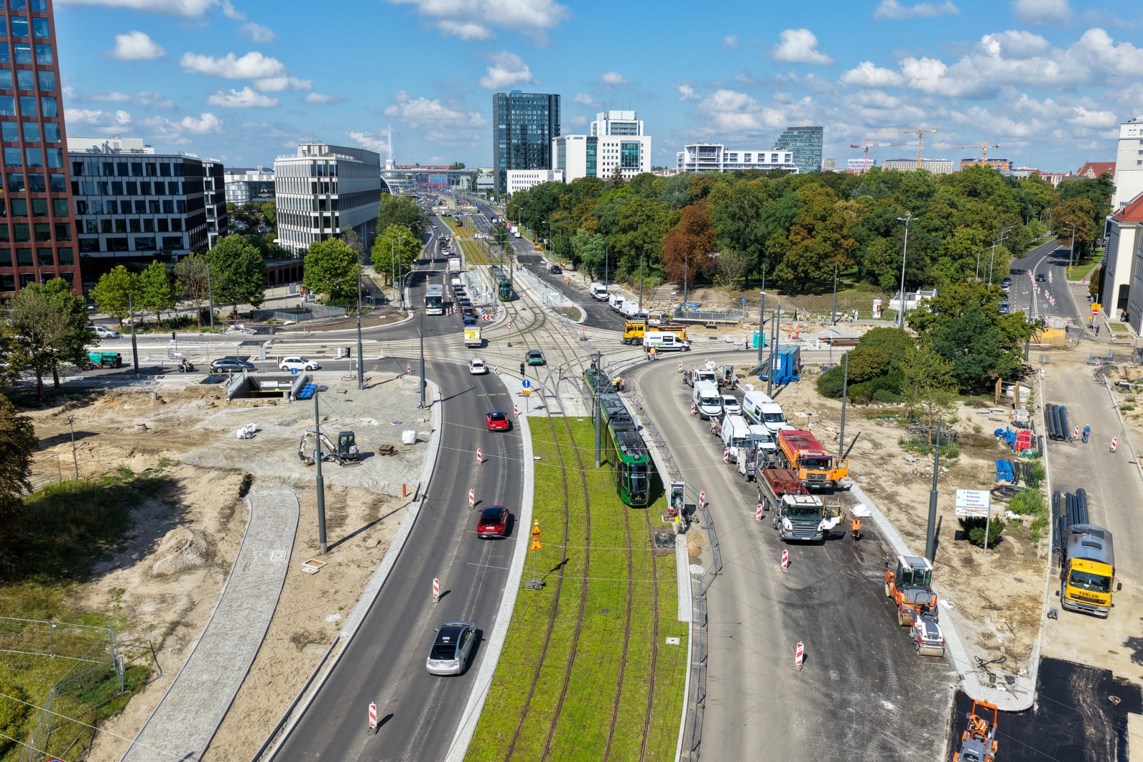 Widok z lotu ptaka na plac budowy. Pośrodku widać torowisko, a na nim jadący tramwaj. W tle samochody, wysokie budynki miejskie i zieleń parku.