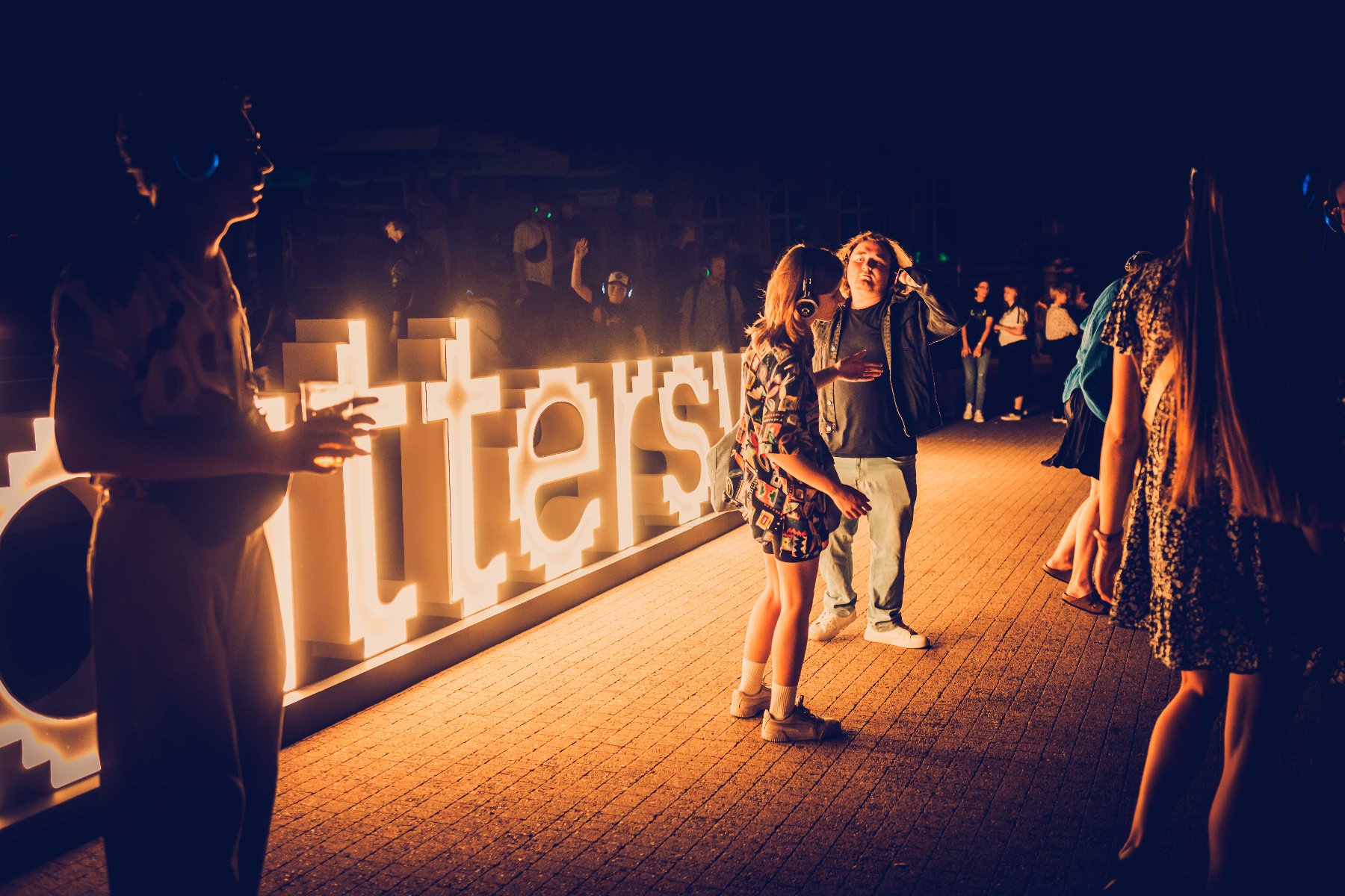Wieczorne wydarzenie, prawdopodobnie koncert, na którym ludzie w słuchawkach tańczą i rozmawiają na świeżym powietrzu. W tle widoczne są podświetlone napisy.