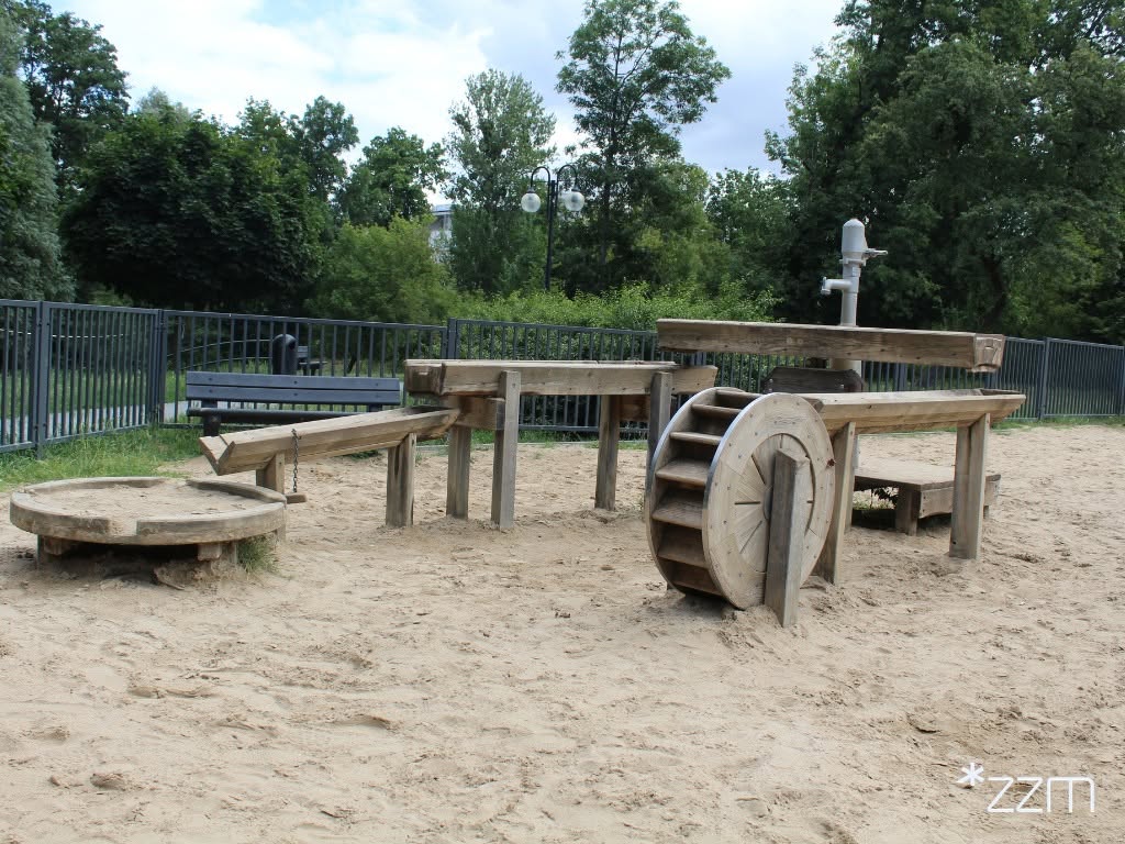 Wodny plac zabaw "Archimedes" na Nadolniku - drewniane koryta, pompa i koło wodne na piaszczystym podłożu.