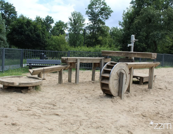 Wodny plac zabaw "Archimedes" na Nadolniku - drewniane koryta, pompa i koło wodne na piaszczystym podłożu.