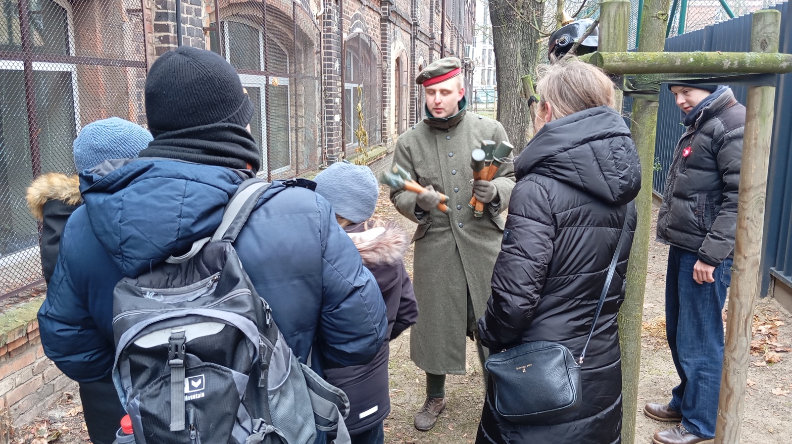 Wokół mężczyzny ubranego w mudur żołnieski z epoki, trzymającego w rękach repliki granatów stoją zgromadzeni ludzi, przypatrując się temu co pokazuje.