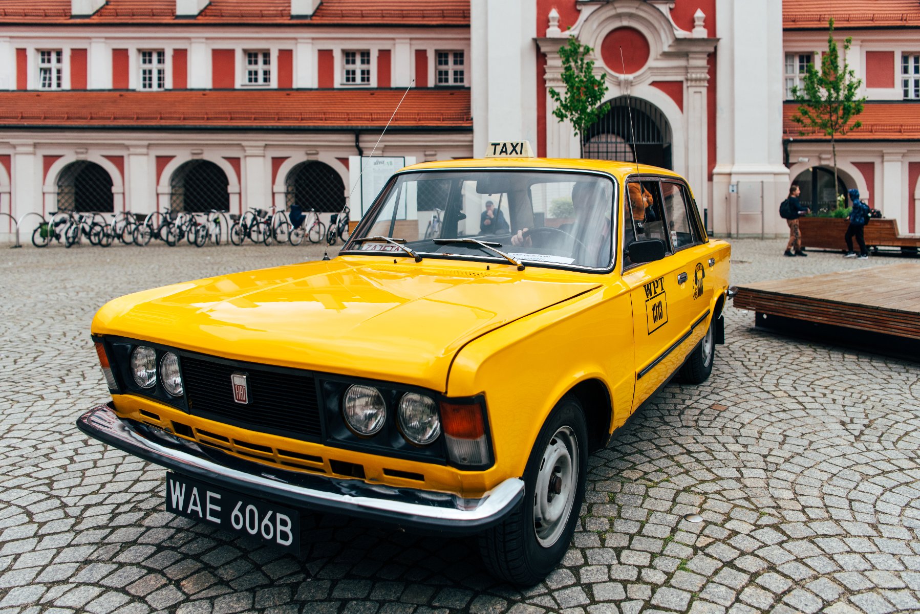 Zabytkowy, żółty Fiat 125p stylizowany na taksówkę Warszawskiego Przedsiębiorstwa Taksówkowego (z numerem boczny WPT 1313 i czarną rejestracją WAE 606B) stoi na brukowanym placu Kolegiackim w Poznaniu. W tle widać charakterystyczny, biało-różowy budynek urzędu miasta (dawne kolegium jezuickie).