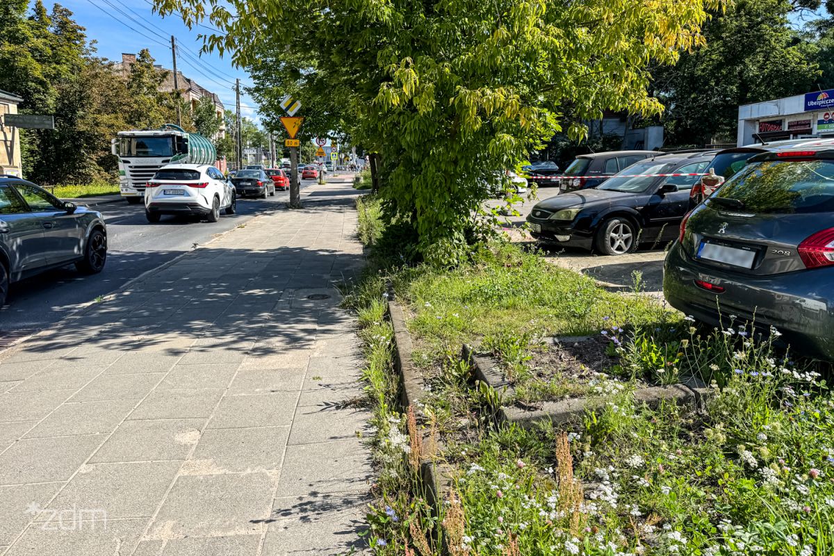 Zdjęcie przedstawia chodnik, jezdnię, po której jeżdżą samochody oraz parking z samochodami, a także pas zieleni.