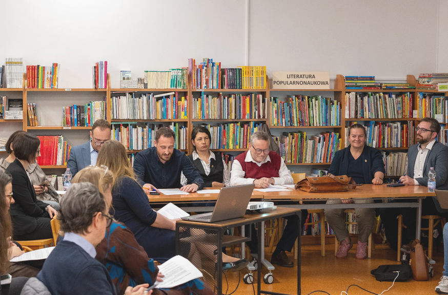 Zdjęcie zostało wykonane w bibliotece. Radni siedzą przy stole, na tle książek. - grafika artykułu